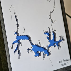 May include: A framed paper art piece depicting Lake Amistad, Del Rio, TX. The lake is cut out of the paper and painted blue, with the surrounding land in gray. The frame is dark gray wood.