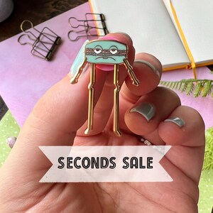 Op de afbeelding: Een gouden speld in de vorm van een macaron met een verdrietig gezicht en de tekst "SECONDS SALE" op een wit lint.