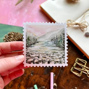 May include: A watercolour painting of a lake scene with mountains in the background. The lake is surrounded by trees and there are white flowers in the foreground. The painting is on a white sticker with a scalloped edge.
