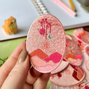 May include: Pink embroidered patch with a crescent moon, a tree, and pink and red mountains. The patch is oval shaped and has a white background with a subtle sparkle effect.