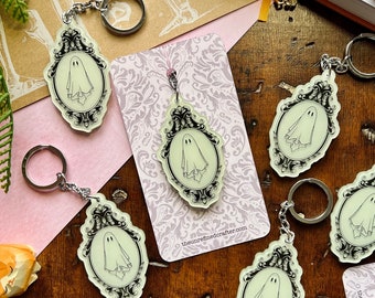 May include: Five glow-in-the-dark keychain charms featuring a ghost in a vintage frame. The charms are made of clear acrylic and have a black outline. The keychains are attached to silver key rings.
