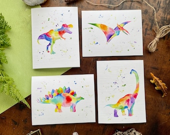 May include: Set of four watercolor dinosaur cards with rainbow colored dinosaurs on a white background with colorful splatter paint. The dinosaurs are a T-Rex, a Pterodactyl, a Stegosaurus, and a Brachiosaurus.