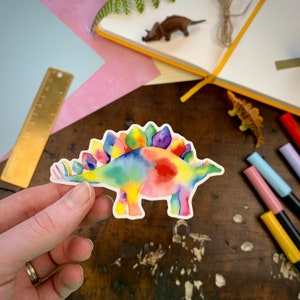 May include: A rainbow tie-dye stegosaurus dinosaur sticker. The sticker is white with a rainbow watercolor design.