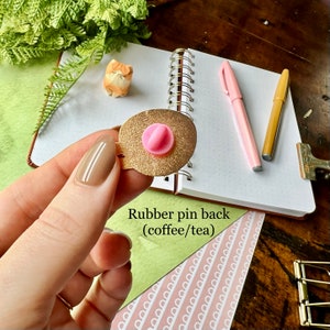 May include: Close-up of the back of a gold-colored enamel pin with a pink rubber pin back. The text "Rubber pin back (coffee/tea)" is visible below the pin.