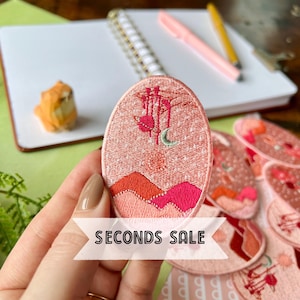 May include: A pink embroidered patch with a crescent moon, stars, and a mountain range. The patch is oval shaped and has a white background with pink and red embroidery. The text "SECONDS SALE" is printed in white on a black banner.