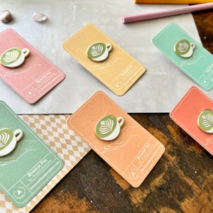 May include: Six enamel pins featuring a green and white latte art design on a white cup. The pins are displayed on cards with the text "Enamel Pin by: Krystal Gonzalez theunrefinedcrafter.com".