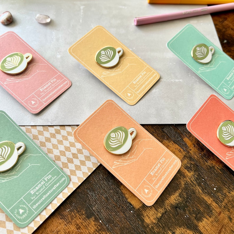 May include: Six enamel pins featuring a green and white latte art design on a white cup. The pins are displayed on cards with the text "Enamel Pin by: Krystal Gonzalez theunrefinedcrafter.com".