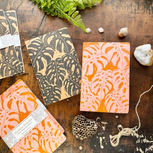 Peut inclure: Quatre cahiers avec un motif de feuilles tropicales. Les cahiers ont des feuilles noires et blanches, et des feuilles roses et orange. Les cahiers sont sur une surface en bois avec un motif en nid d'abeille et un morceau de ficelle.