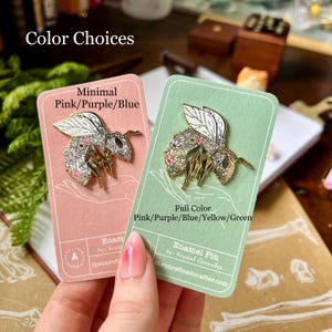 May include: Two cards showing different color options for a gold enamel pin of a bee with floral details. The card on the left shows a minimal color scheme with pink, purple, and blue. The card on the right shows a full color scheme with pink, purple, blue, yellow, and green. The text on the cards reads "Minimal Pink/Purple/Blue" and "Full Color Pink/Purple/Blue/Yellow/Green".