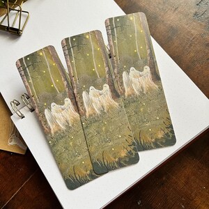 May include: Three bookmarks featuring a forest scene with glowing lights and white ghosts. The bookmarks are printed on a textured paper with a green and brown background.