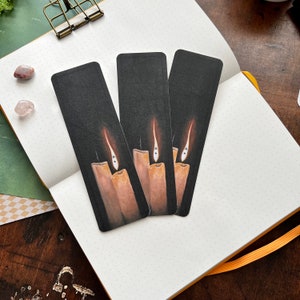 Puede incluir: Tres marcadores negros con un dise&ntilde;o de tres velas con llamas y una cara fantasmal en cada llama. Los marcadores est&aacute;n en un cuaderno blanco punteado con un borde naranja.