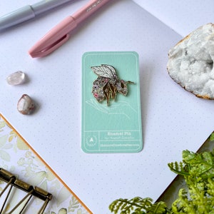 May include: A gold enamel pin featuring a bee covered in pink and white flowers. The pin is on a light blue card with the text "Enamel Pin by Krystal Giestales theunrefinedcrafter.com".