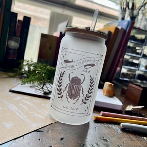 May include: A frosted glass tumbler with a brown wooden lid and a straw. The tumbler has a graphic of a beetle with the text "Beetle Collector's Club est. 2019".