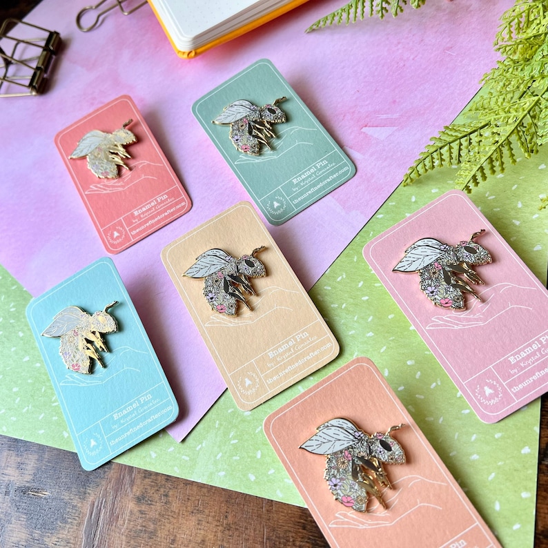 May include: Five enamel pins featuring a bee with floral details on a white background. The pins are displayed on cards with the text "Enamel Pin" and the website address "theunrefine.etsy.com".