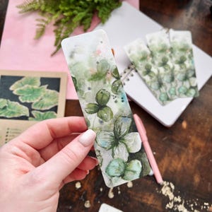 May include: A set of three watercolor bookmarks featuring a green and white floral design. The bookmarks are printed on a white background and have a slightly textured surface.