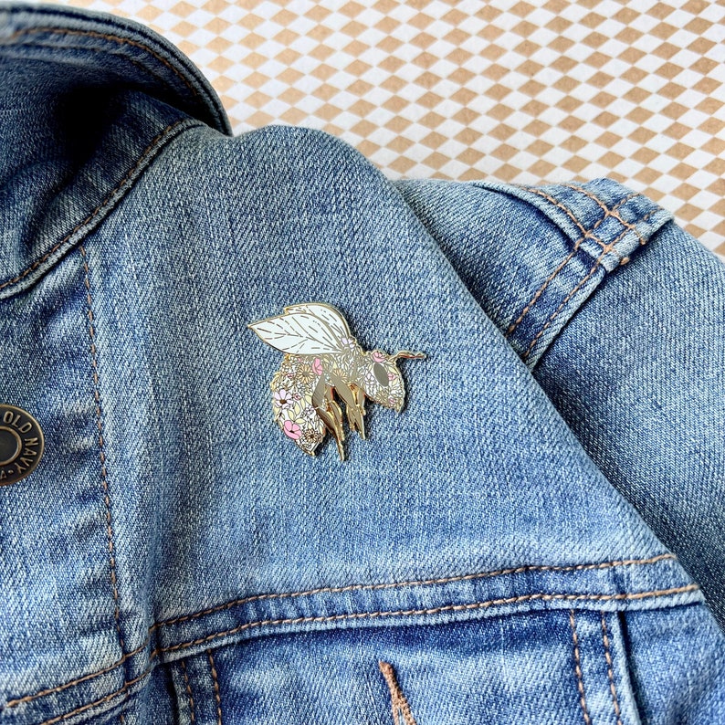 May include: A gold bee pin with white wings and pink and yellow floral details. The pin is on a blue denim jacket.
