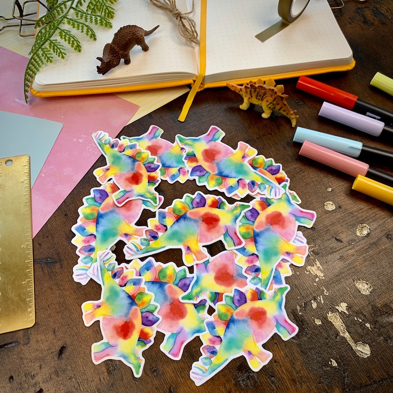 May include: A set of nine colorful dinosaur stickers with a rainbow tie-dye pattern. The stickers are made of a glossy vinyl material and are perfect for adding a fun and whimsical touch to any project.