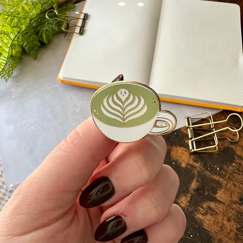 May include: Gold-tone enamel pin of a white cup with a green matcha latte and latte art. The pin has a white background and a gold outline.
