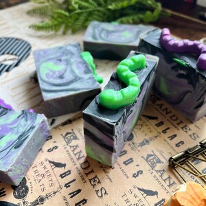 Pode incluir: Quatro barras de sabonete feitas à mão com tema de Halloween. As barras são pretas e verdes com redemoinhos de cor e um verme verde no topo. As barras de sabonete estão sobre uma superfície marrom com um fundo de papel vintage.