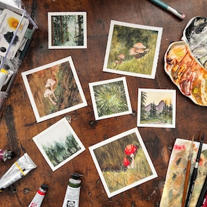 Può includere: Una collezione di piccoli dipinti ad acquerello raffiguranti scene di boschi e funghi. I dipinti presentano vari colori, tra cui verdi, marroni e rossi. Materiali artistici, tra cui tubetti di vernice, pennelli e una tavolozza, sono sparsi intorno ai dipinti su una superficie di legno.