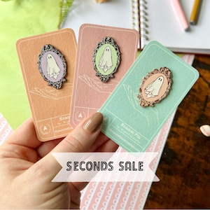 May include: Three enamel pins featuring a ghost in a frame. The pins are on cards with a light green, light pink, and peach background. The cards have text that reads "Enamel Pin by The Spotted Owl Inc. thespottedowlart.com".