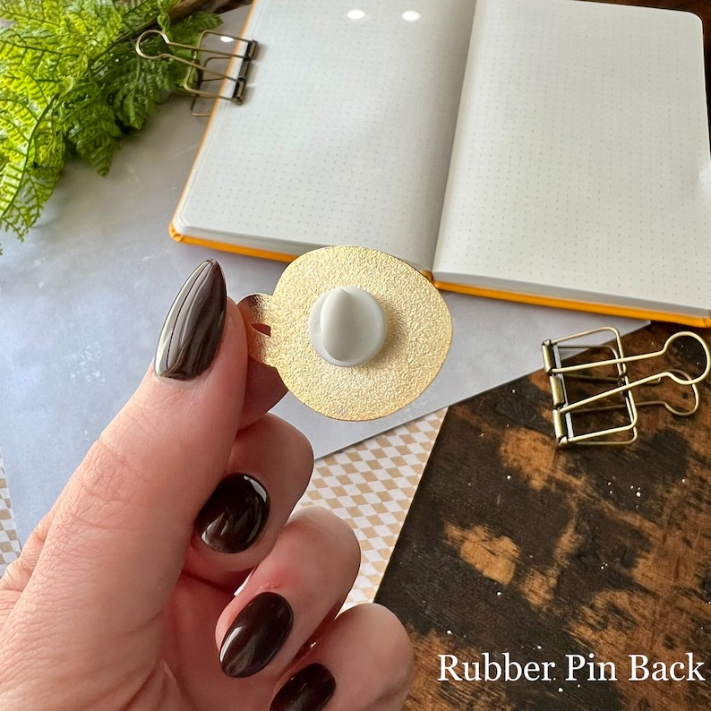 May include: Close-up of the back of a gold-colored pin with a white rubber pin back. The text "Rubber Pin Back" is visible below the pin.