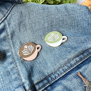 May include: Two enamel pins shaped like coffee mugs with latte art. One mug is white with green latte art and the other is pink with brown latte art. The pins are on a blue denim jacket.