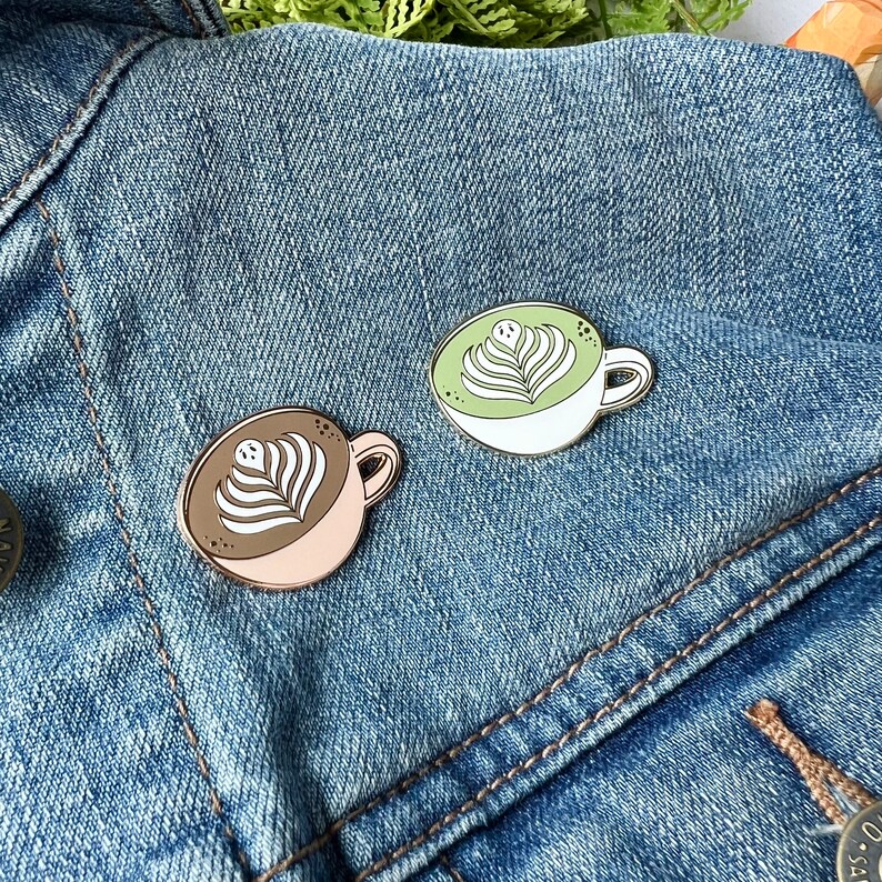 May include: Two enamel pins shaped like coffee mugs with latte art. One mug is white with green latte art and the other is pink with brown latte art. The pins are on a blue denim jacket.
