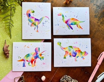 May include: Set of four watercolor dinosaur cards with rainbow colors and white splatter paint backgrounds. The dinosaurs are a velociraptor, a plesiosaur, a triceratops, and an ankylosaurus.