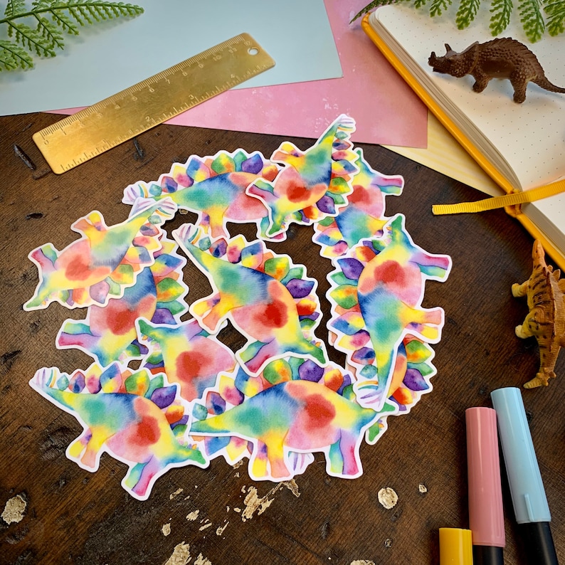 May include: A set of colorful rainbow tie-dye dinosaur stickers. The dinosaurs are all facing the same direction and have a white outline.