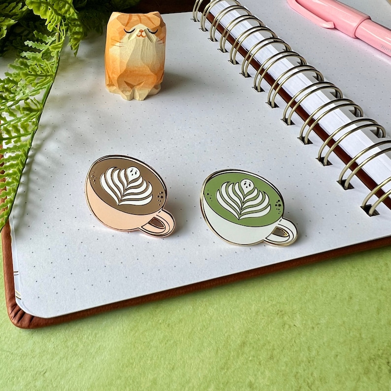 May include: Two enamel pins depicting a cup of coffee and a cup of matcha latte. Both cups have a white foam design with a ghost face in the center.