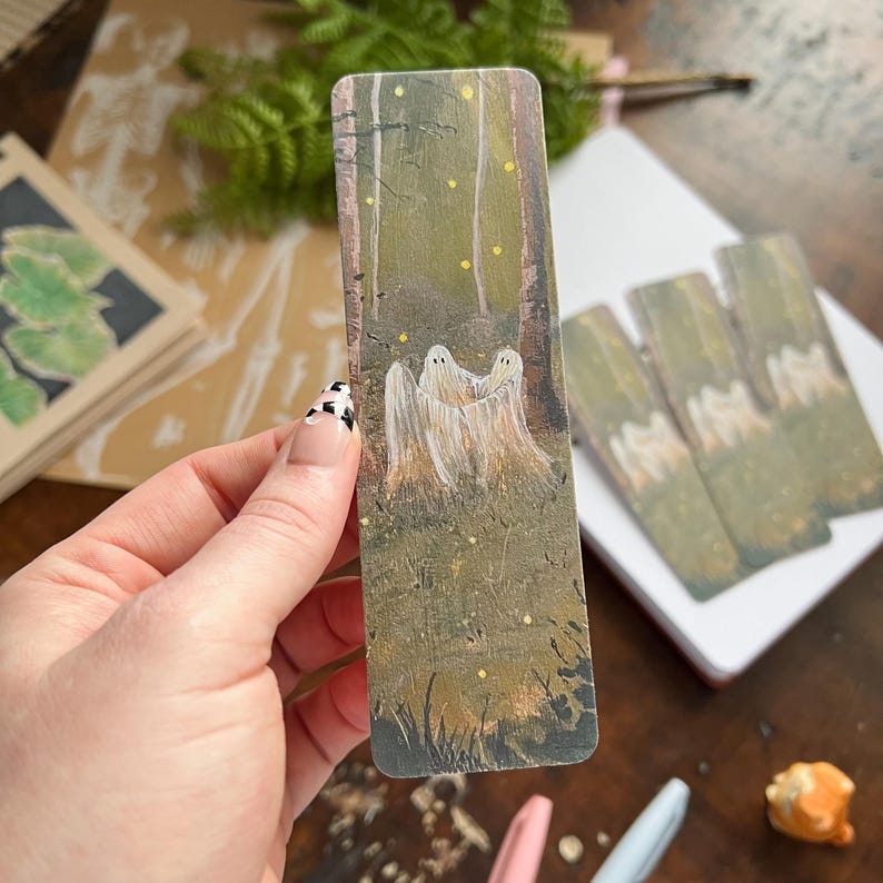 May include: A hand holds a bookmark with a painted image of three ghosts in a forest. The ghosts are white and translucent, and the forest is green and brown. The background is a blurry image of a wooden table.