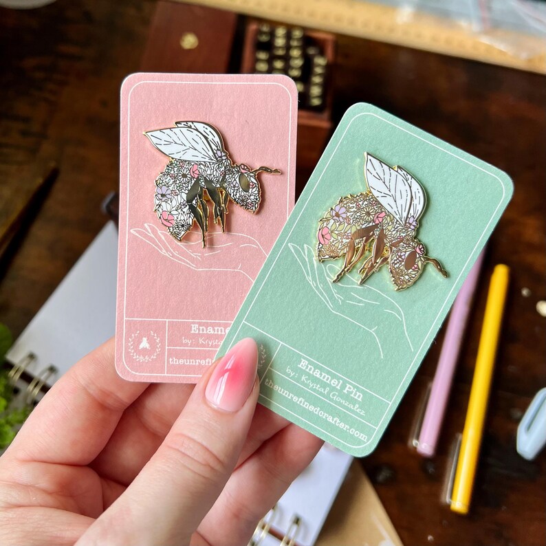 May include: Two enamel pins featuring a bee with floral details. The pins are displayed on pink and green cards with the text "Enamel Pin by Krystal Gonzalez theunrefinedcrafter.com".