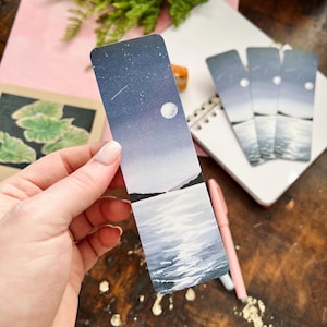 May include: A watercolour bookmark depicting a night sky scene. The bookmark features a crescent moon, stars, and a shooting star over a dark blue body of water. The water reflects the light of the moon.