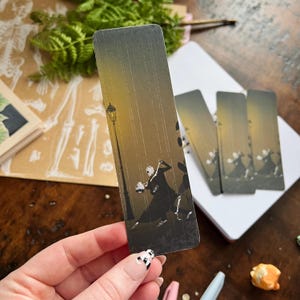 May include: A black and white bookmark featuring a skeleton couple dancing under a street lamp in the rain. The background is a muted yellow and brown.
