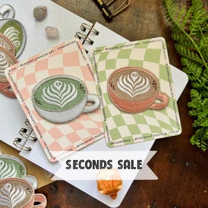 May include: Two embroidered patches of latte art in mugs. The patches are on a pink and green checkered background. The text "SECONDS SALE" is in the foreground.