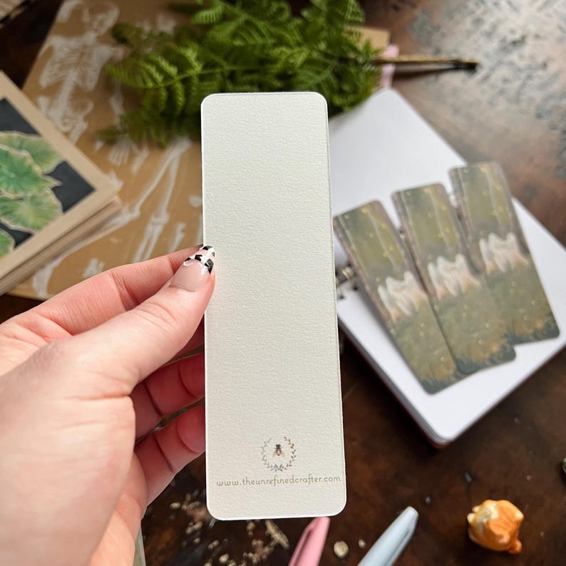 May include: A white bookmark with a textured surface. The bookmark has a small bee logo and the text "www.theunrefinedcrafter.com" printed on the bottom.