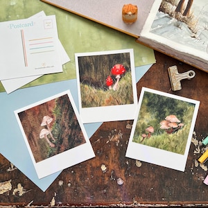 Może przedstawiać: Trzy akwarelowe wydruki artystyczne z ilustracjami grzybów. Wydruki są w stylu polaroid z białymi obramowaniami. Widoczna jest również pocztówka z kolorowymi liniami i napisem "Postcard". Tło obejmuje drewnianą powierzchnię i materiały artystyczne.