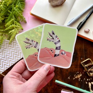 May include: Two stickers featuring a black and white striped creature with red eyes emerging from a pink sand dune. The creature has a large mouth with sharp teeth. The stickers are on a wooden surface.