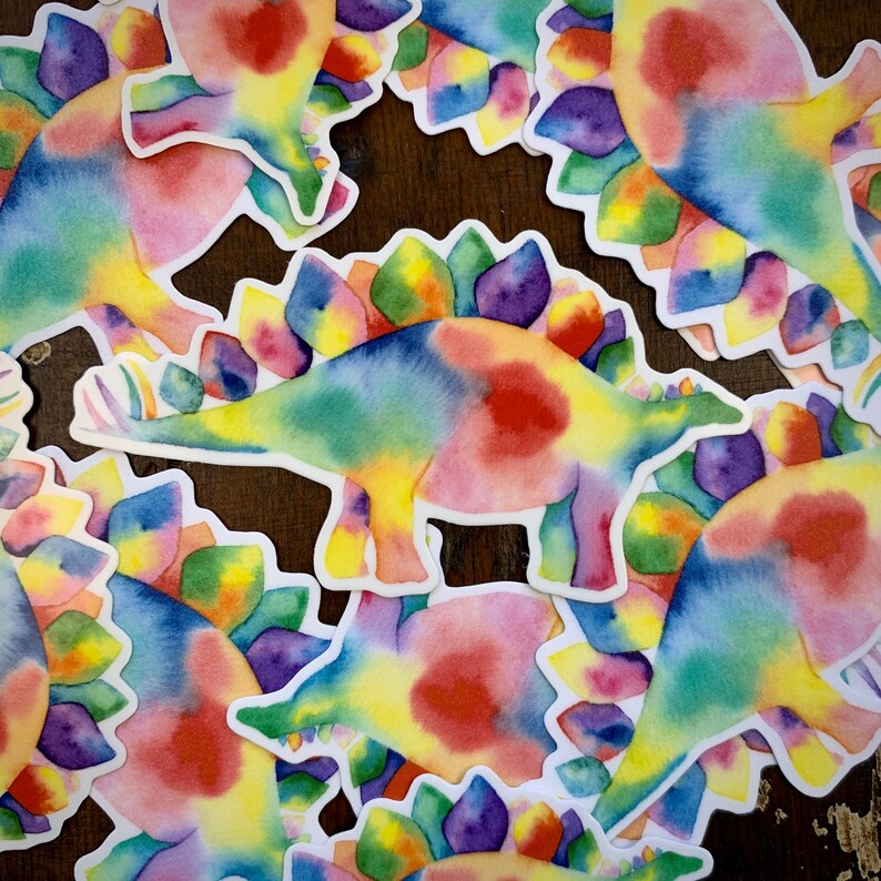May include: A set of colorful dinosaur stickers with a rainbow watercolor design. The stickers feature a variety of dinosaurs, including a stegosaurus, a triceratops, and a brachiosaurus.