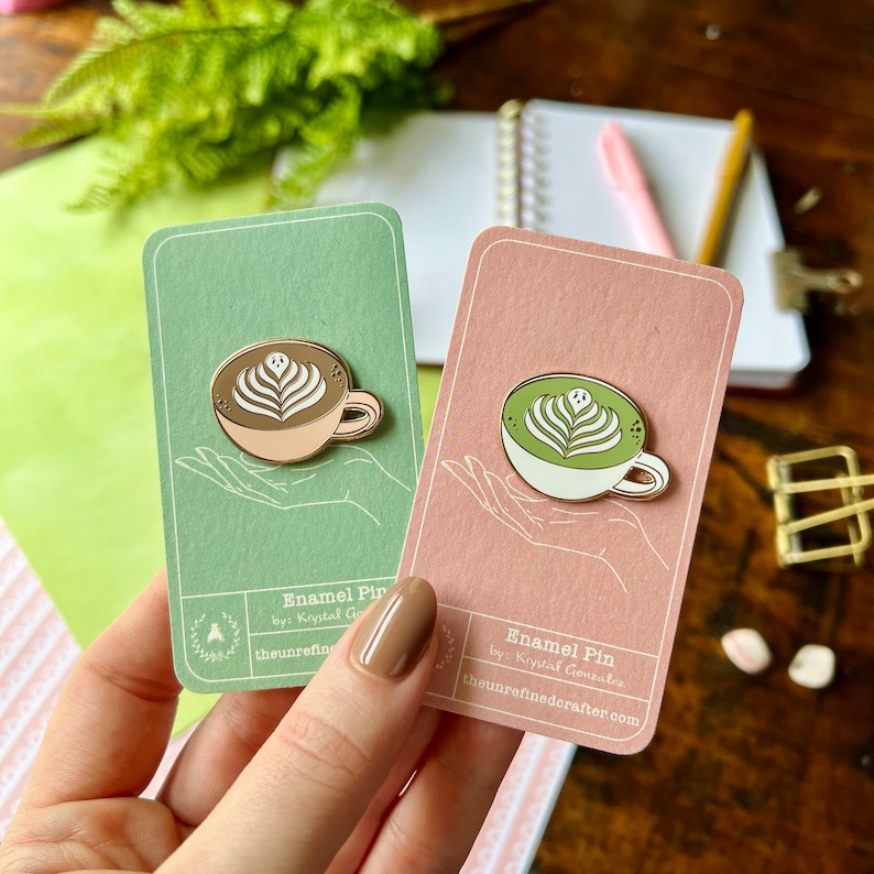 May include: Two enamel pins featuring a latte and a matcha latte in a white cup with a gold rim. The latte art is a heart shape with a swirl on top. The pins are on a green and pink card with the text "Enamel Pin by: Krystal Gonzalez theunrefinedcrafter.com".