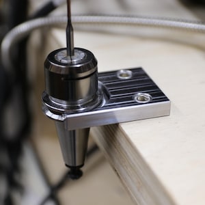 May include: Close-up of a silver metal CNC machine tool holder with a cutting bit inserted. The tool holder is mounted on a silver metal base with two screw holes. The background is blurred.