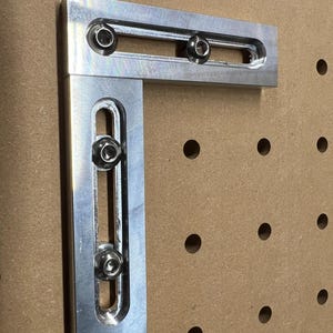 May include: A right-angle metal bracket with three bolts. The bracket is made of silver-colored metal and has a rectangular shape with rounded edges. The bolts are silver and secure the two perpendicular sections of the bracket.