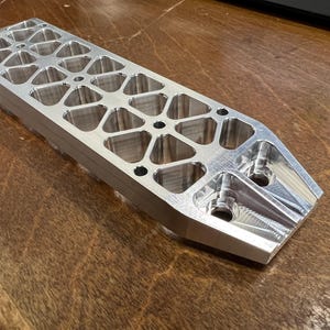 Könnte beinhalten: Ein rechteckiges, silberfarbenes Metallbauteil mit einer Reihe von dreieckigen Aussparungen und Bohrungen. Das Bauteil hat eine polierte Oberfläche und liegt auf einer Holzoberfläche. Das Design deutet auf ein Präzisionsteil hin.
