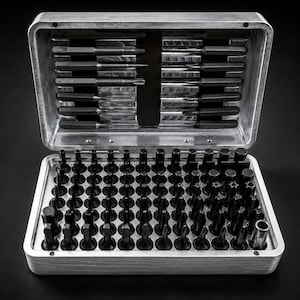 Op de afbeelding: Een zilverkleurige metalen opbergkoffer met een grote collectie zwarte schroevendraaierbits en metalen gereedschappen. De koffer staat open en toont rijen van verschillende bitvormen en maten die in een raster zijn gerangschikt.