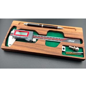 May include: A wooden case containing a digital caliper, a pen, and small metal tools. The caliper features a red digital display, and the brand name "HAAS" is visible. The case is made of dark wood with green felt inserts.