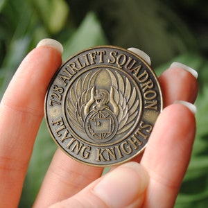 May include: A bronze-colored coin with the inscription "728 Airlift Squadron Flying Knights" and a winged emblem.