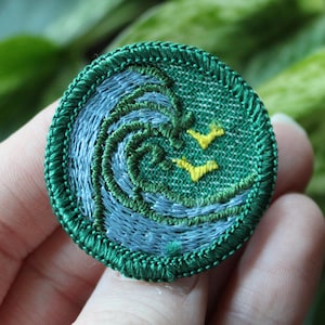 May include: A round, embroidered patch featuring a stylized ocean wave design in shades of blue and green. Two yellow birds are embroidered above the wave. The patch has a green border and is held in a hand.