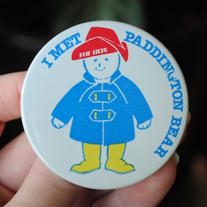 Może przedstawiać: Okrągła, biała przypinka z napisem „I MET PADDINGTON BEAR” na obrzeżach. Przedstawia ilustrację misia Paddington w niebieskim płaszczu i czerwonym kapeluszu. Klasyczna postać dla dzieci.