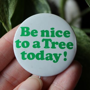 May include: A white button with the text "Be nice to a Tree today!" in green. The button is held in a hand, with a green leafy background.